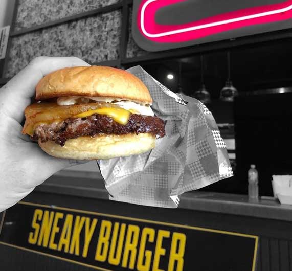 A hand holding a fresh Sneaky burger over the counter, neon Sneaky Burger sign behind